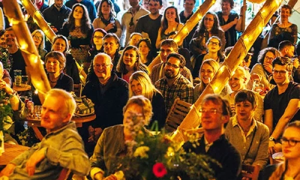 A lively audience gathered in a cozy venue, enjoying a performance under warm lights, featuring a diverse group of men and women engaged and smiling.