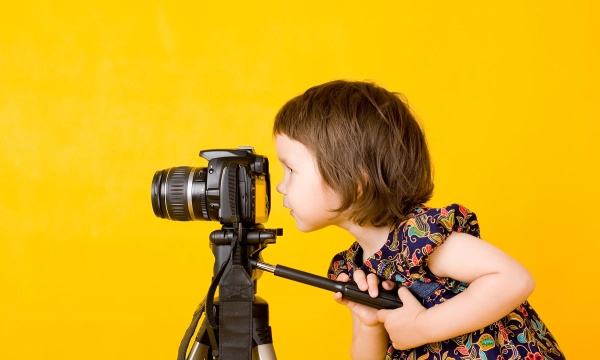 A young girl with short hair in a colorful dress peers closely at a camera mounted on a tripod against a bright yellow background.