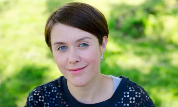 Smiling individual with brown hair and blue eyes, wearing a dark patterned shirt, poses outdoors against a green background.