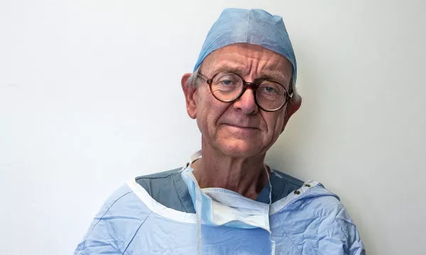 An elderly male doctor wearing a surgical cap, glasses, and a medical gown, looking directly at the camera with a neutral expression against a plain background.