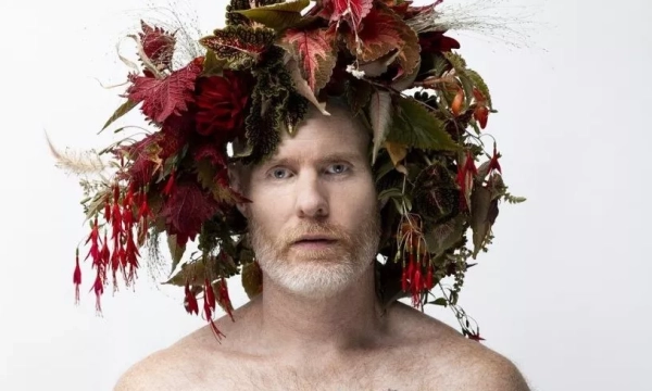 A man with a pale complexion and a beard wears a crown made of colorful leaves and flowers, posing against a plain white background.