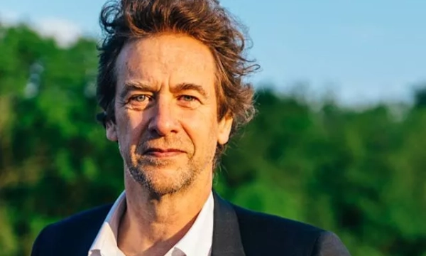 A man with tousled hair stands outdoors against a backdrop of greenery, wearing a dark blazer and a white shirt, looking confidently at the camera.