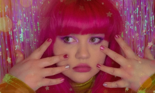 A person with vibrant pink hair and colorful nails poses in front of a sparkling, starry backdrop, expressing a whimsical, playful aesthetic.