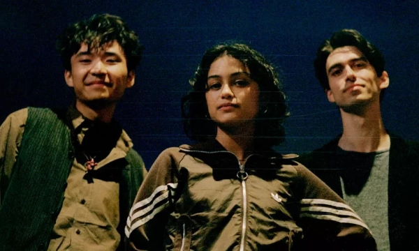 Three young adults stand against a dark backdrop, exuding confidence, with two men flanking a woman in a sporty jacket.