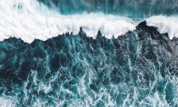 Aerial view of turbulent ocean waves crashing on a dark blue sea, creating frothy white foam and a textured surface.