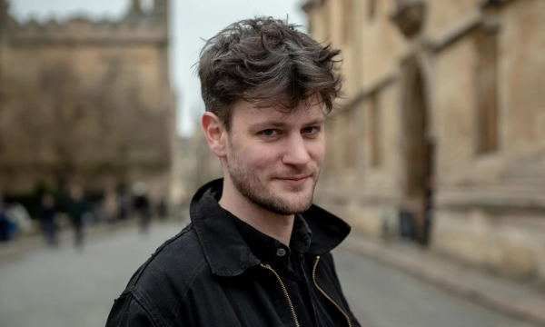 A young man with tousled hair stands confidently on a street lined with historic buildings, hinting at a university setting.