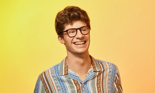 A smiling young man wearing glasses and a colorful patterned shirt against a bright yellow and orange gradient background.