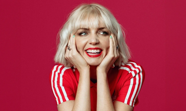 A smiling woman with short blonde hair and red lipstick, wearing a red sports jersey with white stripes, against a bright pink background.