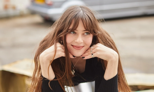 A woman with long, wavy hair poses outdoors, resting her chin on her hands and smiling softly.