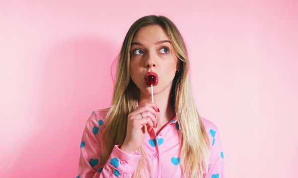 A young woman in a pink, heart-patterned shirt holds a red lollipop, looking pensively at the camera against a pink background.