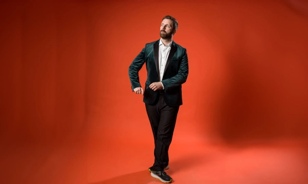 A man in a green suit jacket and black pants poses against an orange backdrop, looking thoughtfully at his watch.