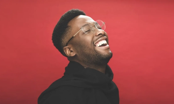 A smiling man with glasses and short hair, wearing a black hooded garment, against a bright red background.