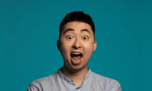 A young man with short black hair and an excited expression, arms crossed, against a solid turquoise background.