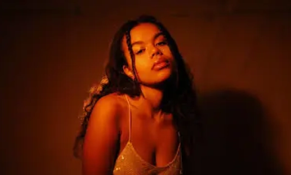 A young woman with curly hair poses in warm, soft lighting, wearing a sparkling spaghetti strap top. Her expression is confident and contemplative.
