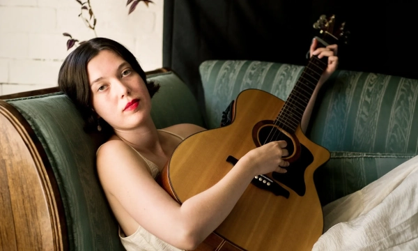 A young woman with short dark hair and bright red lips rests on a vintage green sofa, holding an acoustic guitar, exuding a relaxed and artistic vibe.