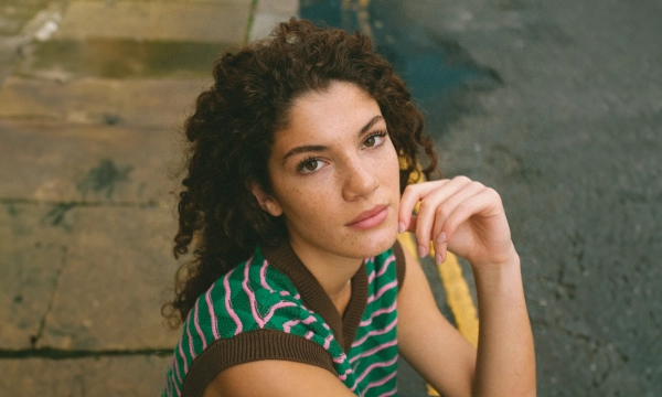 A young woman with curly hair and freckles sits casually on a street, resting her chin on her hand, wearing a striped sweater.