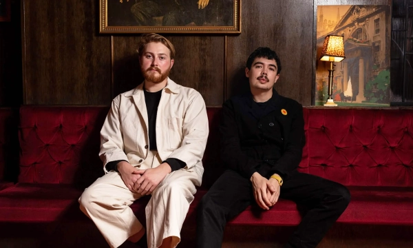 Two young men sit side by side on a red couch in a dimly lit room adorned with vintage decor.