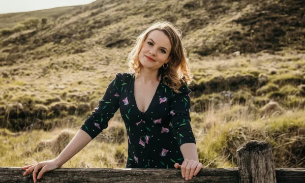 A woman in a floral dress leans against a wooden fence, smiling warmly against a scenic, grassy landscape.