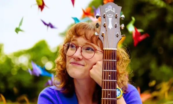A young woman with curly hair and glasses rests her chin on a guitar, smiling thoughtfully amidst colorful paper cranes in the background.
