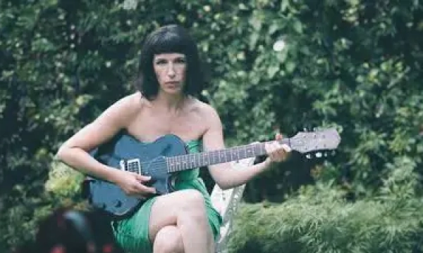 A woman in a green dress sits outdoors, playing a blue guitar, surrounded by lush greenery.