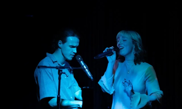A male and female singer perform on stage, illuminated by blue lighting, with the male playing an instrument and the female singing into a microphone.