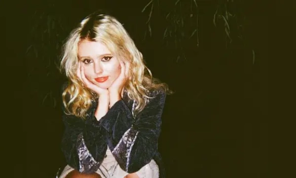 A blonde young woman with wavy hair sits with her hands on her cheeks, wearing a dark jacket and looking directly at the camera.