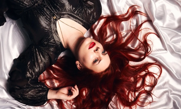 A woman with long red hair lies on a white satin surface, wearing a dark, shimmery dress and bold red lipstick, gazing upward.