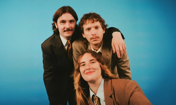 Three individuals in suits pose against a bright blue background, displaying friendly expressions. Their hairstyles and attire reflect a vintage style.