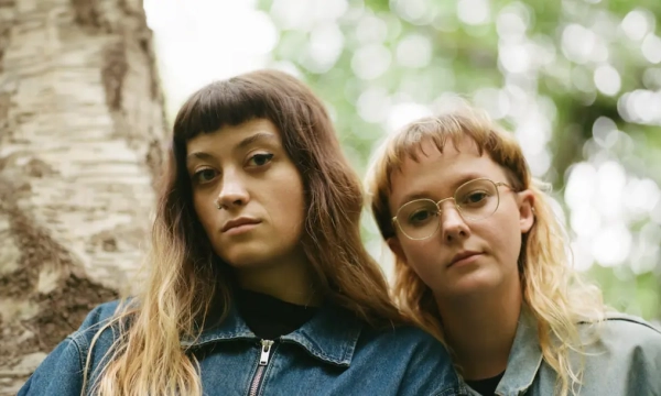 Two young women with long hair, one wearing glasses, pose against a blurred green background of trees, dressed casually in denim jackets.