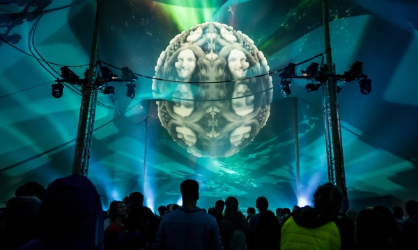A large illuminated spherical projection displays abstract visuals under a vibrant tent, while a crowd gathers to watch the immersive experience.