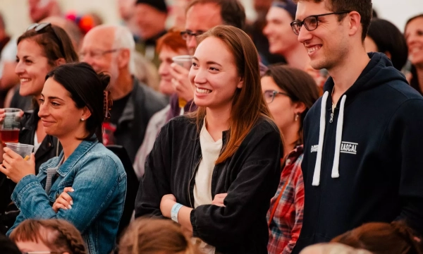 A diverse group of smiling people enjoying a live event, with focus on a woman in a black jacket and a man in a hoodie, surrounded by an engaged audience.
