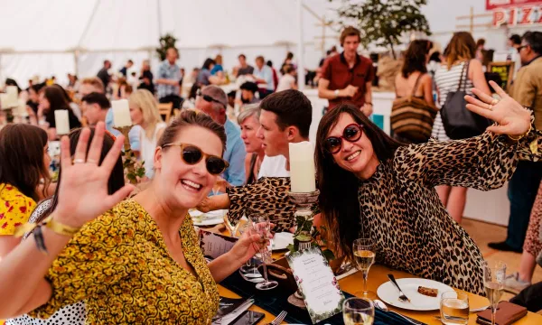Two women in colorful outfits and sunglasses wave cheerfully at the camera while seated at a long dining table in a lively tented setting.