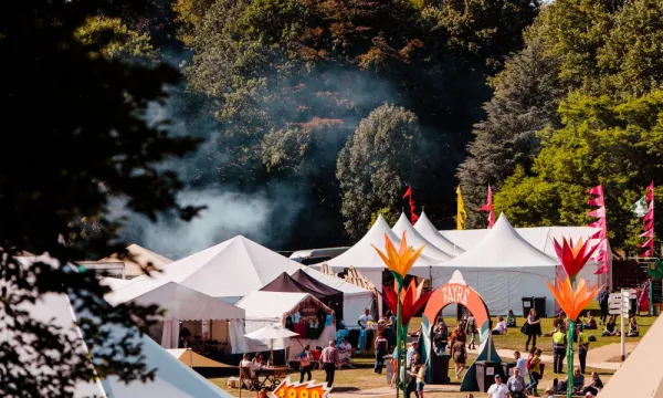 A vibrant outdoor festival scene featuring colorful tents, large flower decorations, and attendees enjoying food and activities in a lush green park setting.