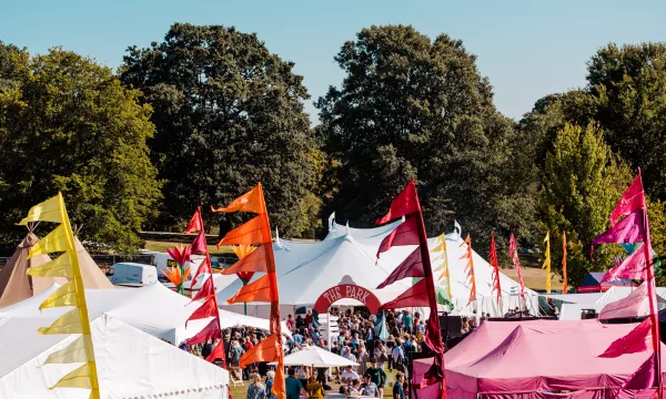 A vibrant outdoor festival scene featuring colorful flags and tents, with large trees in the background and a crowd enjoying the festivities.