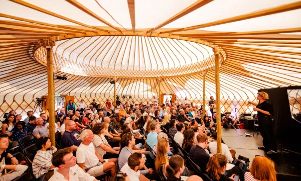 An audience gathered inside a yurt, attentively listening to a speaker on stage, with natural light illuminating the wooden structure.