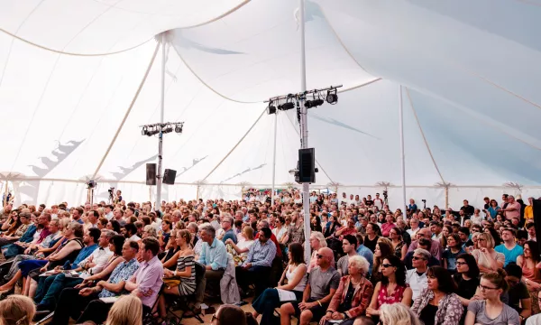 A large audience seated inside a tent during an event, with bright lighting and a vibrant atmosphere.