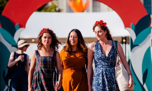 Three women walk under a colorful arch labeled "FAYRE," with floral crowns and festive outfits, enjoying a lively outdoor event on a sunny day.