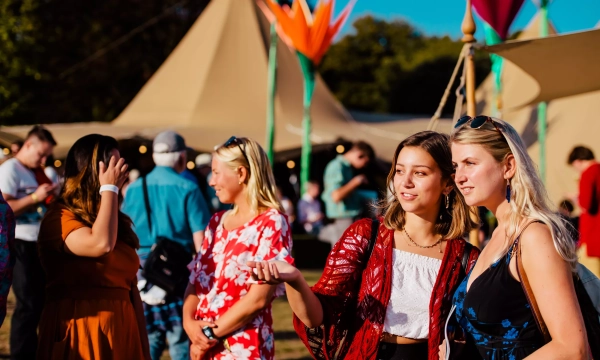 Two young women conversing in a lively outdoor festival setting, surrounded by colorful tents and people enjoying the event.