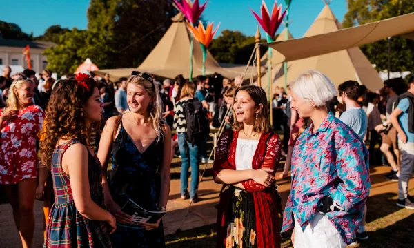 A lively outdoor festival scene with a group of four young women chatting, colorful tents in the background, and a crowd enjoying the event.