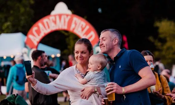 A family takes a selfie at an outdoor event, with festival lights in the background and people enjoying the atmosphere.