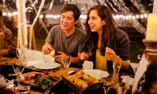 A man and a woman enjoy a meal at an outdoor gathering, seated at a table set with food and drinks under a clear tent with string lights.