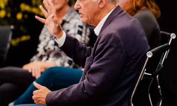 A man in a suit gestures while speaking at a discussion panel, with two women sitting behind him in a partially blurred setting.