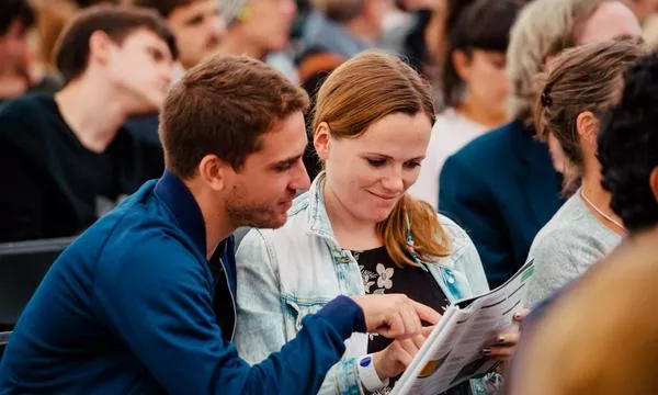A young man and woman sit closely together, reviewing a document while surrounded by an attentive audience in a casual setting.