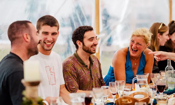 A group of four people laughing and enjoying a meal at a banquet table, surrounded by glasses and plates, in a festive tent setting.