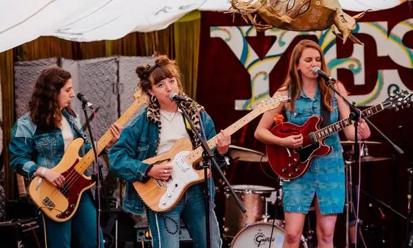 Three female musicians perform on stage, wearing denim outfits, playing instruments including a guitar and a bass, with a colorful backdrop and drums behind them.