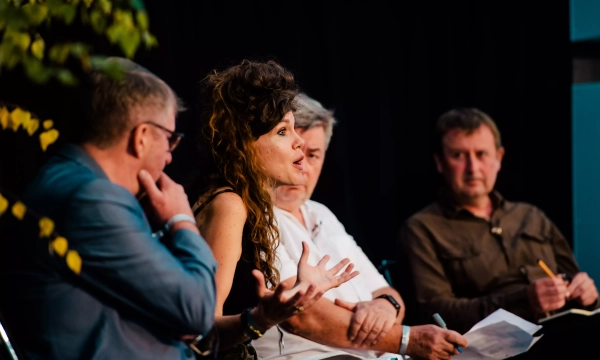 A panel discussion featuring three men and a woman, animatedly speaking, with notes and a plant in the background.