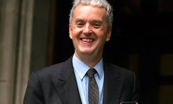 A smiling man in a suit stands outside a building, holding a document, with short gray hair and a light blue shirt.