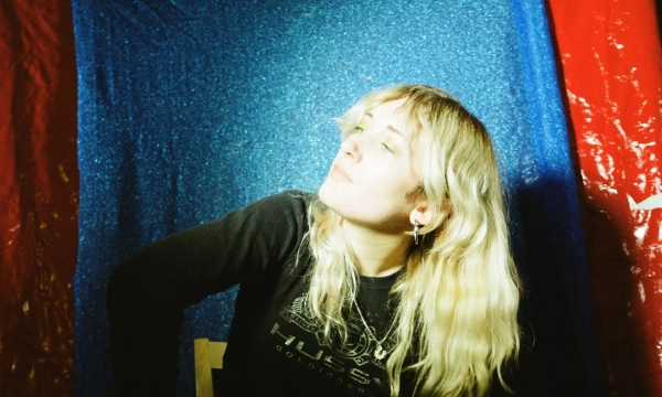 A young woman with long, wavy blonde hair poses against a shimmering blue and red backdrop, wearing a black long-sleeved shirt.