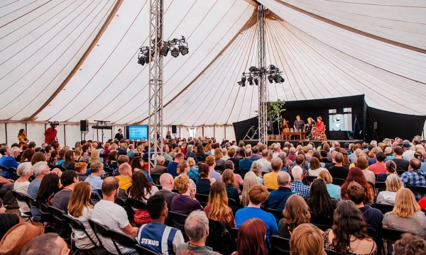 A large audience sits inside a tent, attentively watching a stage presentation with lighting and decorations, creating a vibrant atmosphere.