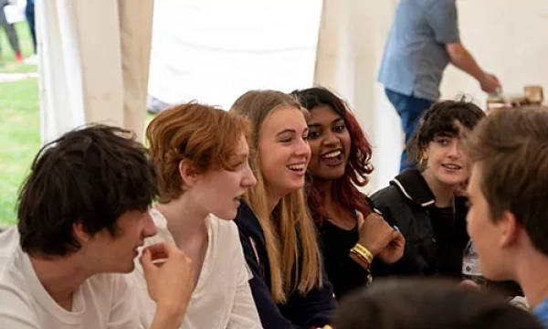 A diverse group of young people sitting together in a tent, laughing and enjoying a conversation, with some people in the background.